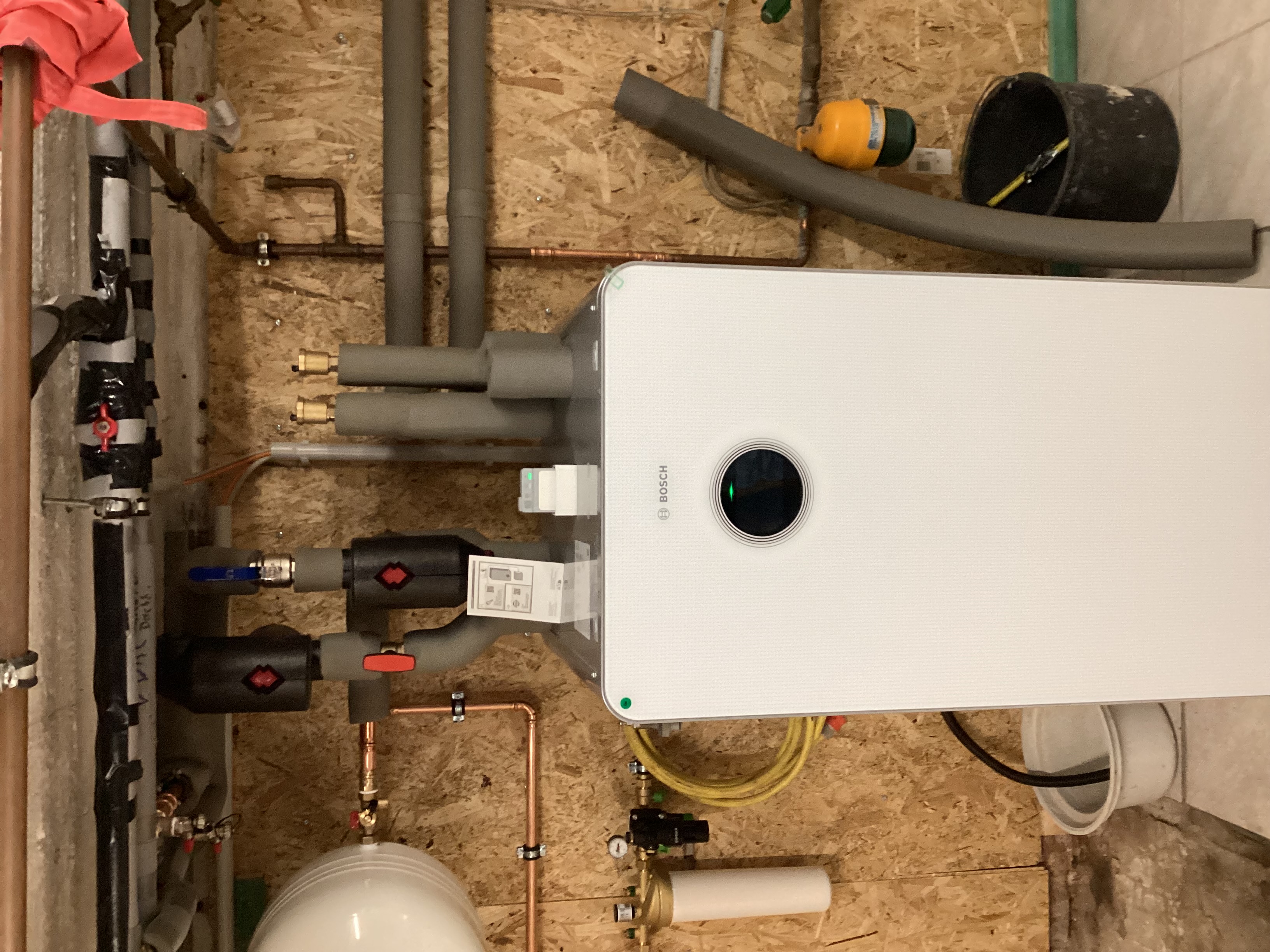 Eine weiße Bosch-Inneneinheit einer Wärmepumpe ist an einer OSB-Holzwand installiert, umgeben von gedämmten Leitungen und Technik.